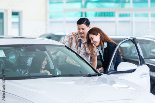 Beautiful young couple choosing a car at the dealership talking to the salon manager in a new car at the dealership professional salesman showing the automobile characteristics buying cars. © tong2530
