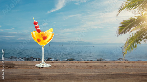 Travel. Summer bright and tasty refreshing cocktail with sea or ocean beach with palms and white sand on background. Yellow alcohol drink with berries. Concept of summertime, vacation, resort, chill.