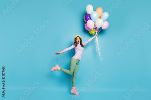 Full length photo of crazy pretty lady hold many air balloons shocked raising up with wind wear purple pullover beret cap green trousers boots isolated blue color background