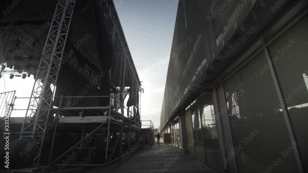 day shot of the back of an enormous music stage , a man walking at narrow passage behind music stage and big building day shot of the back of an enormous music stage , a man walking at narrow passage behind music stage and big building