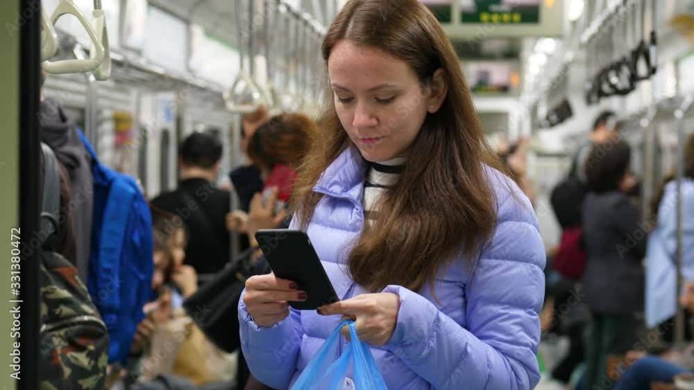 European woman travel by Seoul metro, stare to phone, standing in aisle of moving car. Portrait shot of passenger, blurred background. Train rock and shake during ride