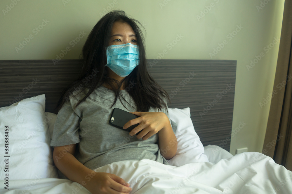 pandemic and virus outbreak - young beautiful scared and worried Asian Chinese woman in medical mask checking online news with mobile phone on bed lockdown at home