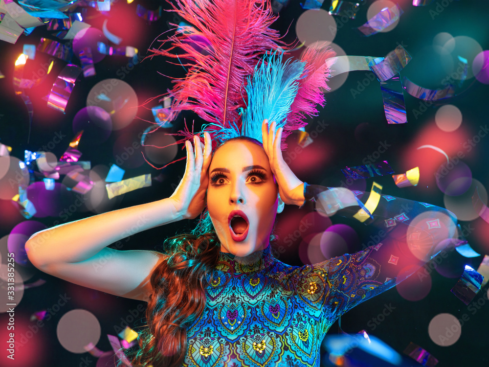 Surprised. Beautiful young woman in carnival, stylish masquerade costume with feathers on black background in neon light, flying confetti. Holidays celebration, dancing, fashion. Festive time, party.