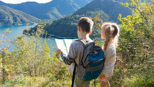 Independent little children scouts with tourist backpacks stand on the shore of a mountain lake in the Alps and pave the way for a family boy scout weekend camping in the forest.