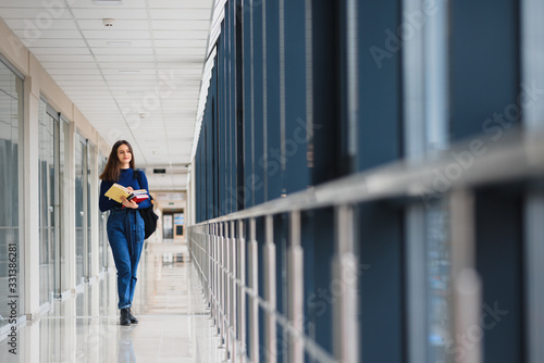Cheerful brunette student girl with black backpack holds books in modern building. female student standing with books in college hallway