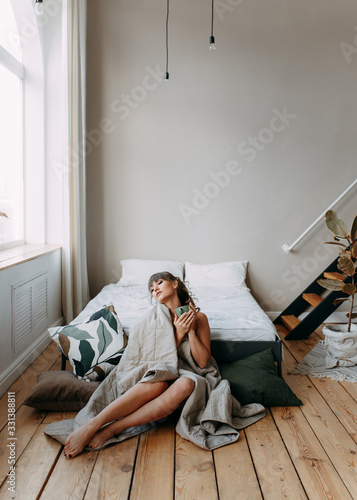 Beautiful happy sweet lonely girl woke up and wrapped in a blanket sitting on the bed in a spacious bright bedroom, resting and drinking coffee, holidays, weekends, hospital regime, cozy home