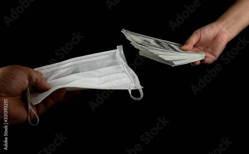 Close up Sell buy medical face mask, Hand holding doctor masks virus protection and money of 100 US dollars banknote a lot of at black background, It costs expensive and high priced products concept