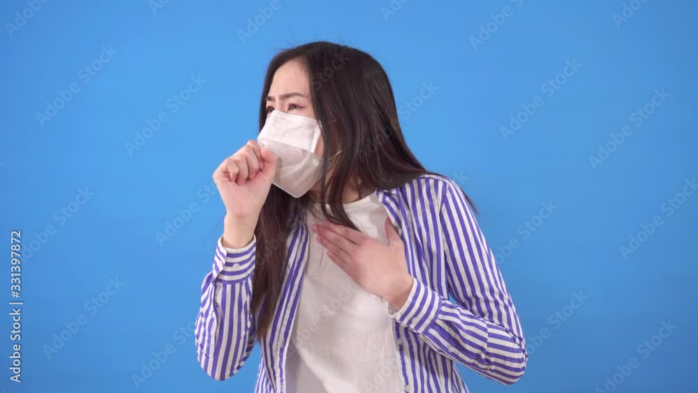 beautiful young asian woman with a protective medical mask on her face coughs while standing on a blue background beautiful young asian woman with a protective medical mask on her face coughs while standing on a blue background