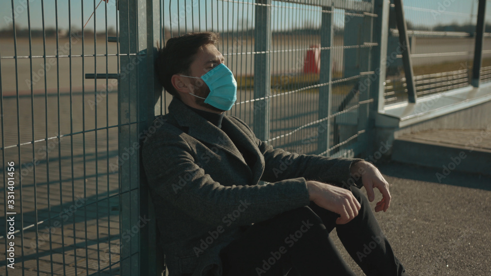 Fototapeta premium Sick man wearing Protective Face Mask sitting near fence border sneeze coughs COVID-19 coronavirus infection pandemic disease virus male tourist epidemic air health illness slow motion
