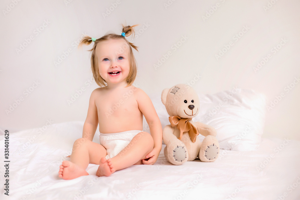 Toddler girl in diapers sits with toy bear on bed at home