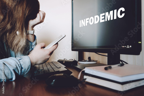 Coronavirus infodemic concept. Frightened woman sitting in front of computer monitor with text infodemic with phone in her hands photo