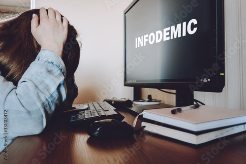 Coronavirus infodemic concept. Frightened woman sitting in front of computer monitor with text infodemic with phone in her hands photo