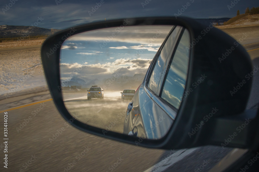 Cars seen in mirror traveling on misty road on transcanadian highway with rocky mountains in the background lit in the late afternoon sun. Epic travel or safety photo.