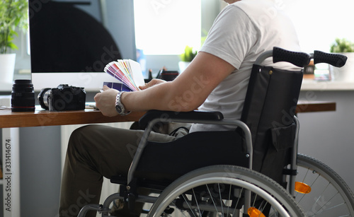 Close-up of professional designer working in office holding colourful palette. Male sitting in wheelchair. Photocamera and laptop on desktop. People with disability concept