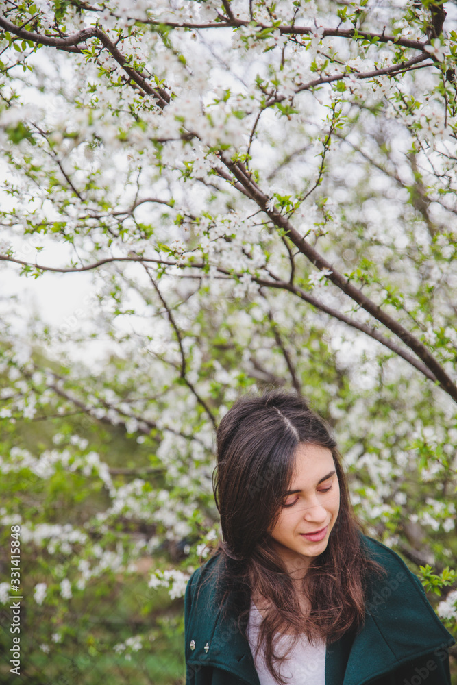 Young caucasian girl with dark hair in white t-shirt and green coat near beautiful white cherry blossom tree