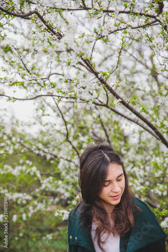 Young caucasian girl with dark hair in white t-shirt and green coat near beautiful white cherry blossom tree