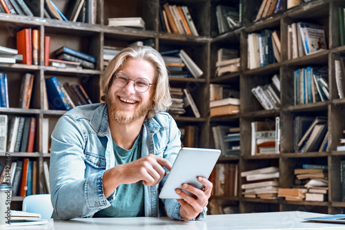 Happy male student teacher smiling laughing holding using digital tablet computer looking at camera e learning easy internet course study online in app concept with device sit at library desk. photo