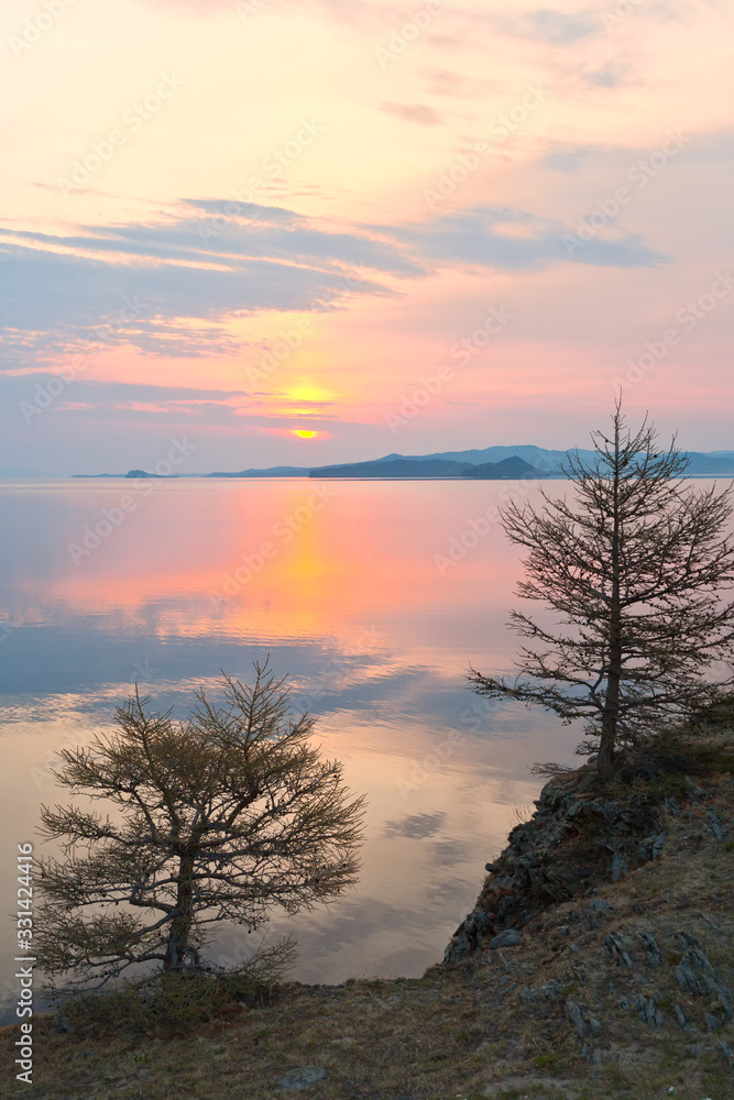 Baikal Lake in the spring at dawn. The rising sun over the Strait of Small Sea. Beautiful quiet landscape with larch trees on the shore in the early morning. Natural background