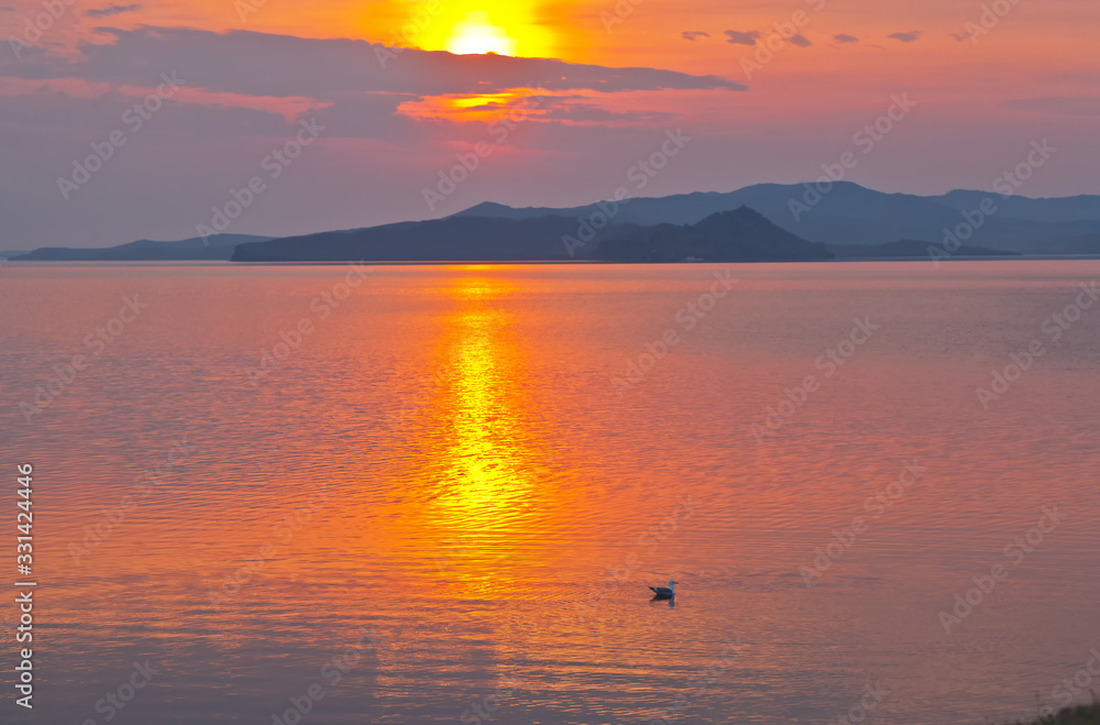 A gorgeous bright colorful sunrise is reflected in the water of Baikal  Lake on an early spring morning. A seagull sways on the waves of the Small Sea. Seascape. Natural background