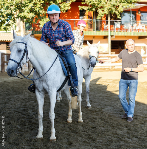 Man in helmet riding by horse at ranch, trainer talking to female