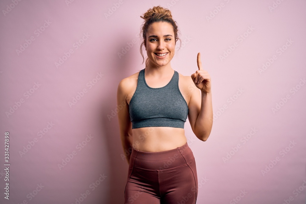 Young beautiful blonde sportswoman doing sport wearing sportswear over pink background showing and pointing up with finger number one while smiling confident and happy.