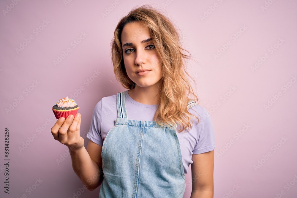 Young beautiful blonde woman eatimg chocolate cupcake over isolated pink background with a confident expression on smart face thinking serious