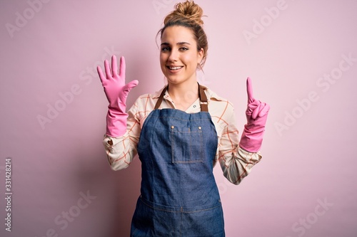 Young beautiful blonde cleaner woman doing housework wearing arpon and gloves showing and pointing up with fingers number six while smiling confident and happy.