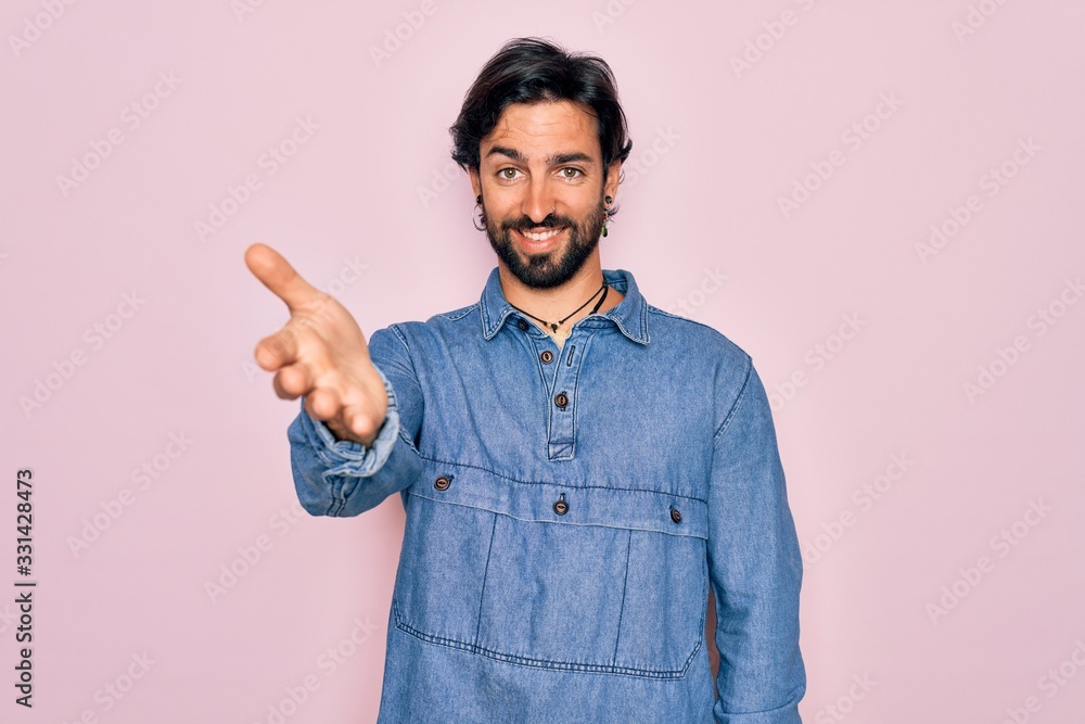 Young handsome hispanic bohemian man wearing hippie style over pink background smiling friendly offering handshake as greeting and welcoming. Successful business. Young handsome hispanic bohemian man wearing hippie style over pink background smiling friendly offering handshake as greeting and welcoming. Successful business.