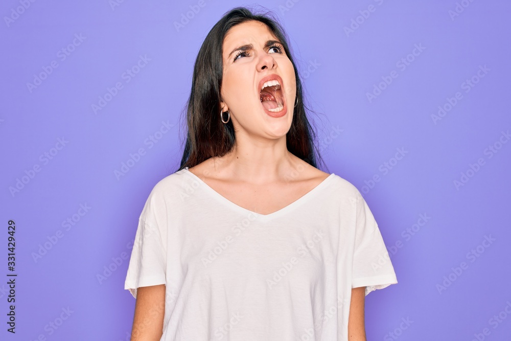 Young beautiful brunette woman wearing casual white t-shirt over purple background angry and mad screaming frustrated and furious, shouting with anger. Rage and aggressive concept.