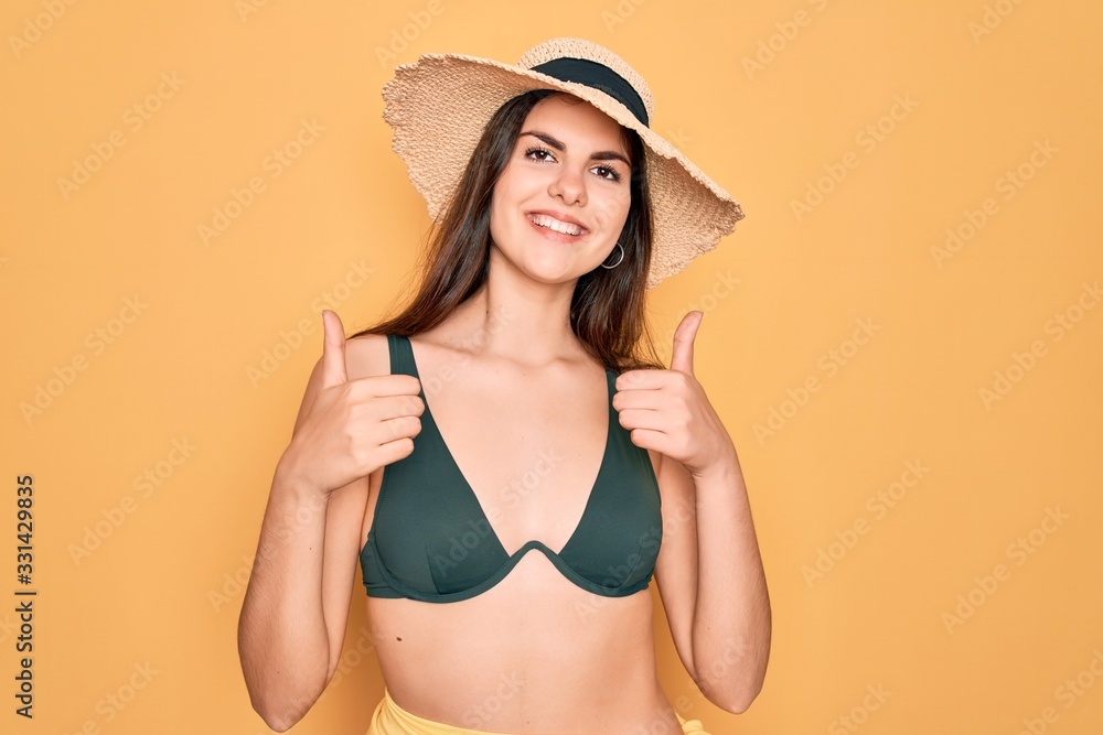 Young beautiful girl wearing swimwear bikini and summer sun hat over yellow background success sign doing positive gesture with hand, thumbs up smiling and happy. Cheerful expression and winner Young beautiful girl wearing swimwear bikini and summer sun hat over yellow background success sign doing positive gesture with hand, thumbs up smiling and happy. Cheerful expression and winner