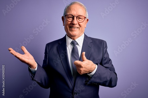 Grey haired senior business man wearing glasses and elegant suit and tie over purple background Showing palm hand and doing ok gesture with thumbs up, smiling happy and cheerful