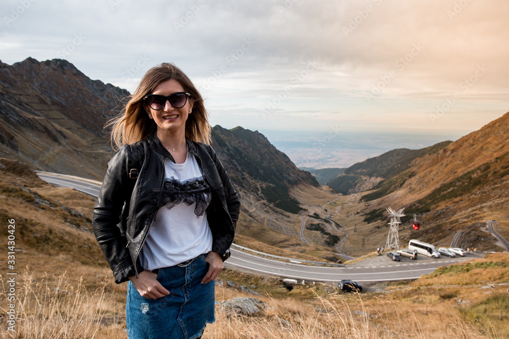 Young beautiful girl or woman portrait standing in a mountain landscape. Travel for fun. Large valley in background with long roads.