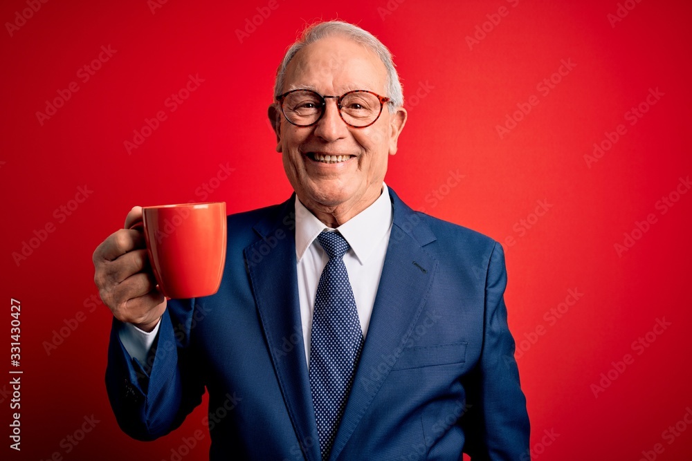 Senior grey haired business man drinking a hot cup of coffee over red background with a happy face standing and smiling with a confident smile showing teeth Senior grey haired business man drinking a hot cup of coffee over red background with a happy face standing and smiling with a confident smile showing teeth