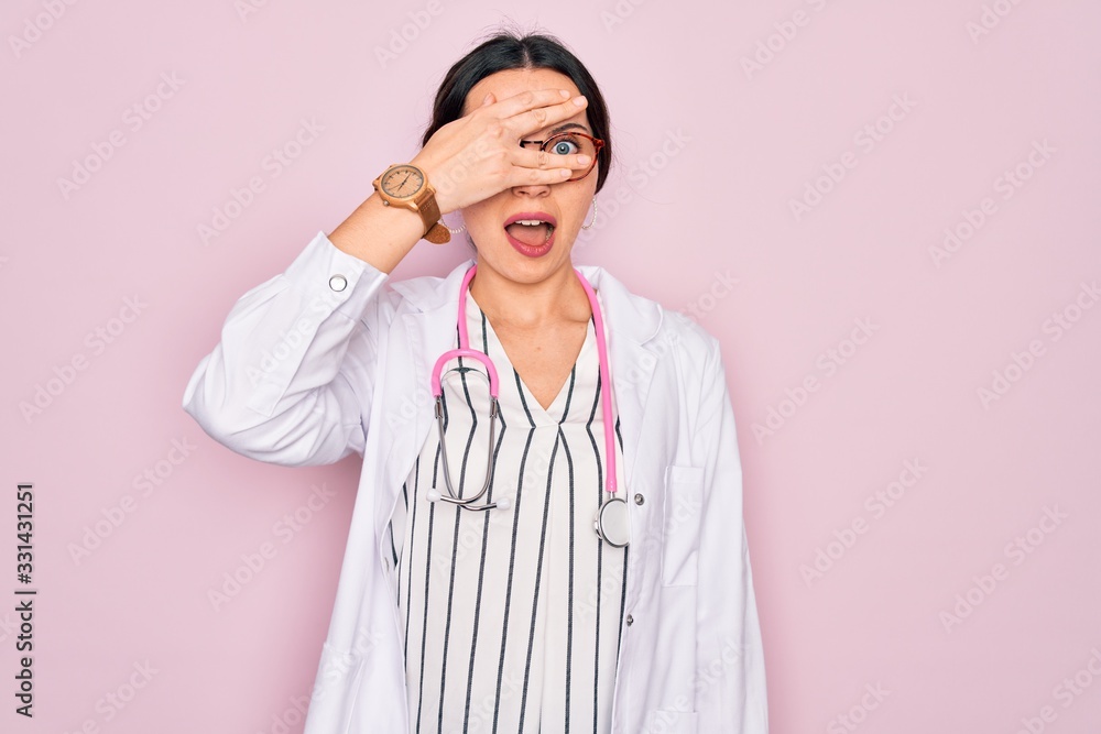 Beautiful doctor woman with blue eyes wearing coat and stethoscope over pink background peeking in shock covering face and eyes with hand, looking through fingers with embarrassed expression. Beautiful doctor woman with blue eyes wearing coat and stethoscope over pink background peeking in shock covering face and eyes with hand, looking through fingers with embarrassed expression.