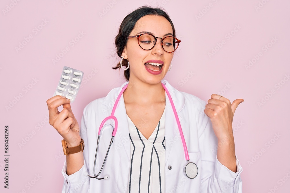 Young beautiful doctor woman with blue eyes wearing stethoscope holding medicine pills pointing and showing with thumb up to the side with happy face smiling Young beautiful doctor woman with blue eyes wearing stethoscope holding medicine pills pointing and showing with thumb up to the side with happy face smiling