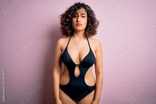 Young beautiful arab woman on vacation wearing swimsuit and sunglasses over pink background Relaxed with serious expression on face. Simple and natural looking at the camera.