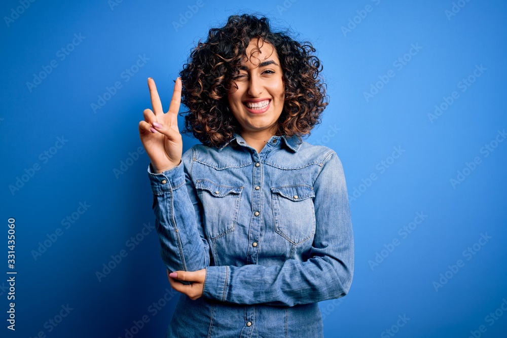 Young beautiful curly arab woman wearing casual denim shirt standing over blue background smiling with happy face winking at the camera doing victory sign. Number two. Young beautiful curly arab woman wearing casual denim shirt standing over blue background smiling with happy face winking at the camera doing victory sign. Number two.