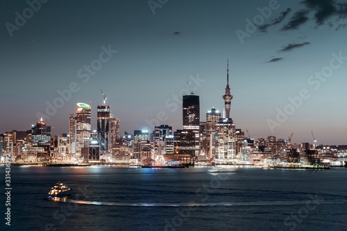 Panorama of Auckland city during blue hour with city lights reflecting in the water and a yacht sailing in the bay, capital of New Zealand