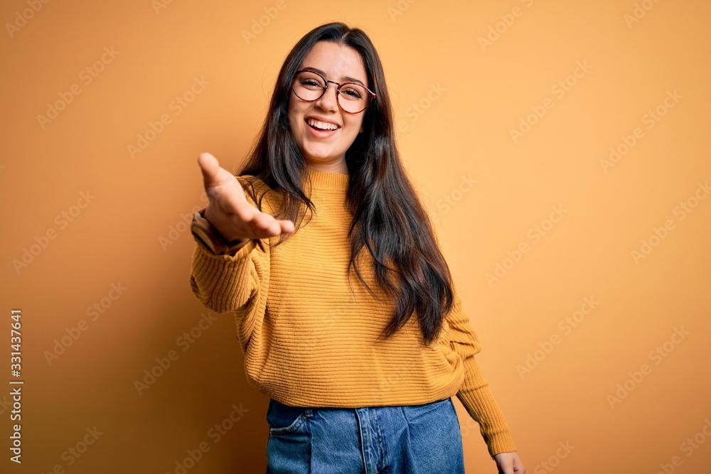 Young brunette woman wearing glasses and casual sweater over yellow isolated background smiling cheerful offering palm hand giving assistance and acceptance. Young brunette woman wearing glasses and casual sweater over yellow isolated background smiling cheerful offering palm hand giving assistance and acceptance.