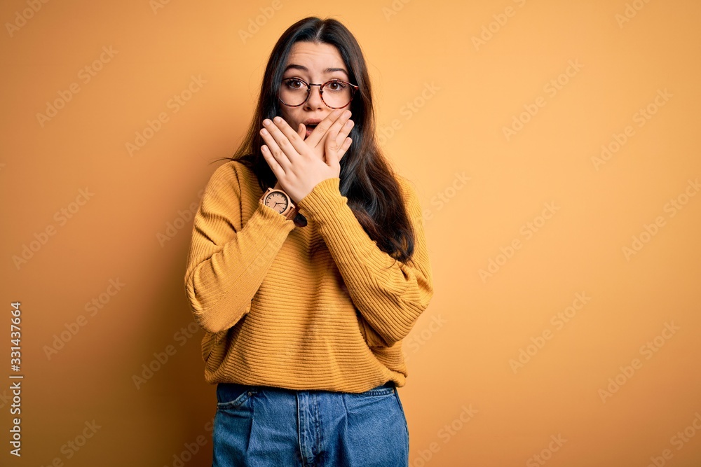 Young brunette woman wearing glasses and casual sweater over yellow isolated background shocked covering mouth with hands for mistake. Secret concept. Young brunette woman wearing glasses and casual sweater over yellow isolated background shocked covering mouth with hands for mistake. Secret concept.