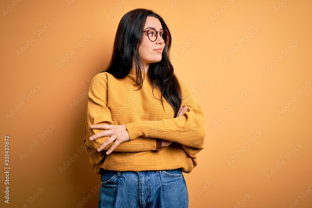 Young brunette woman wearing glasses and casual sweater over yellow isolated background looking to the side with arms crossed convinced and confident Young brunette woman wearing glasses and casual sweater over yellow isolated background looking to the side with arms crossed convinced and confident