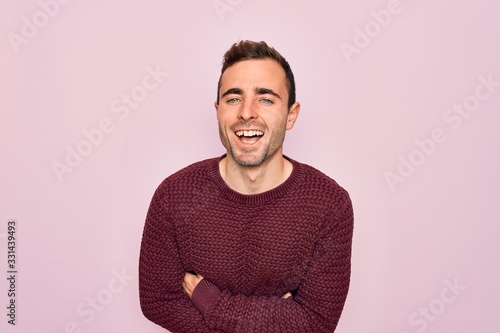 Young handsome man with blue eyes wearing casual sweater standing over pink background smiling and laughing hard out loud because funny crazy joke with hands on body.