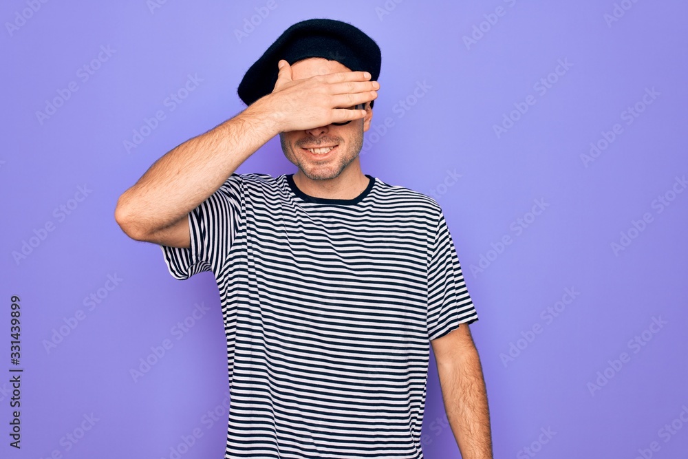 Handsome man with blue eyes wearing striped t-shirt and french beret over purple background smiling and laughing with hand on face covering eyes for surprise. Blind concept. Handsome man with blue eyes wearing striped t-shirt and french beret over purple background smiling and laughing with hand on face covering eyes for surprise. Blind concept.
