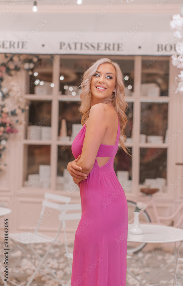 Attractive smiling blonde in pink sexy dress posing among locations stylized as Paris cafe decorated in pink tones with curly flowers