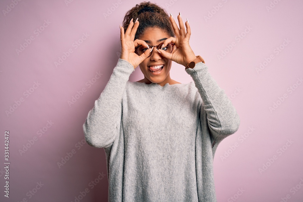 Young beautiful african american girl wearing sweater and glasses over pink background doing ok gesture like binoculars sticking tongue out, eyes looking through fingers. Crazy expression. Young beautiful african american girl wearing sweater and glasses over pink background doing ok gesture like binoculars sticking tongue out, eyes looking through fingers. Crazy expression.