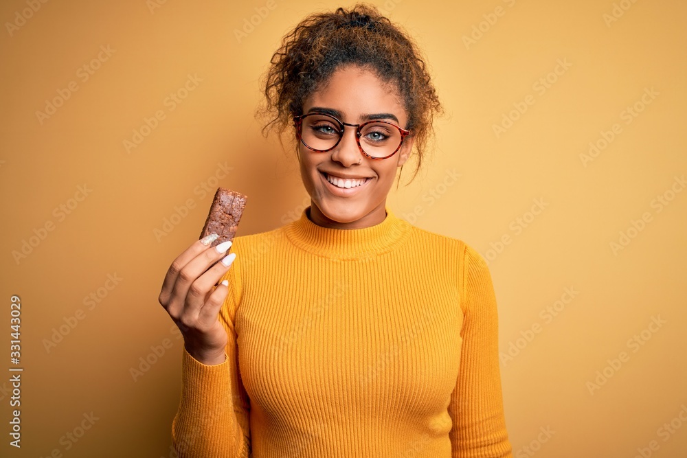 Young african american afro girl eating energetic protein bar of cereal over yellow background with a happy face standing and smiling with a confident smile showing teeth Young african american afro girl eating energetic protein bar of cereal over yellow background with a happy face standing and smiling with a confident smile showing teeth