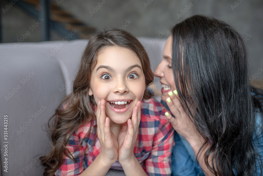 Dark-haired mom telling something interesting to her surprised daughter in the ear Dark-haired mom telling something interesting to her surprised daughter in the ear