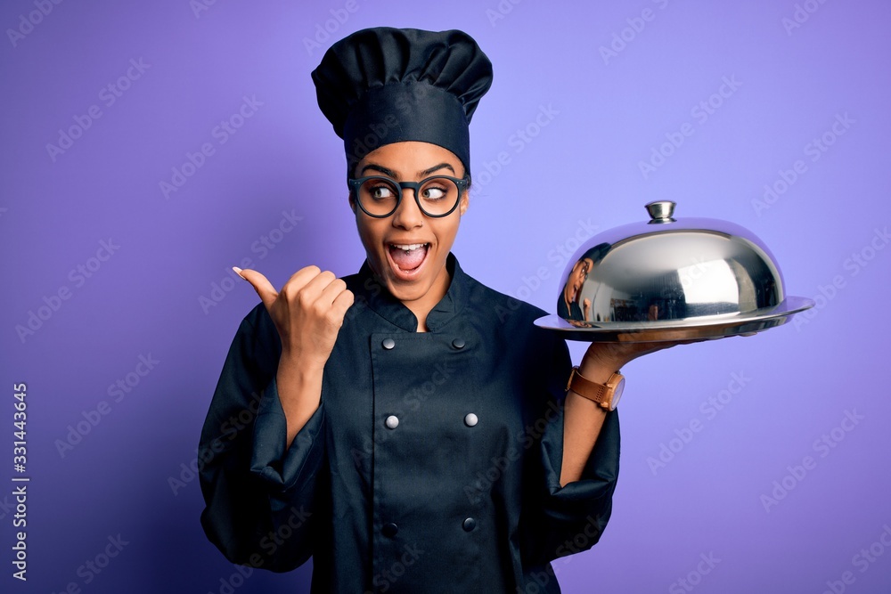 Young african american cooker girl wearing uniform and hat holding tray  with dome pointing and showing with thumb up to the side with happy face  smiling Stock Photo