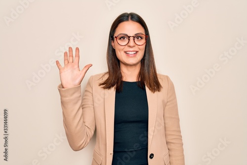 Young beautiful brunette businesswoman wearing jacket and glasses over white background showing and pointing up with fingers number five while smiling confident and happy.
