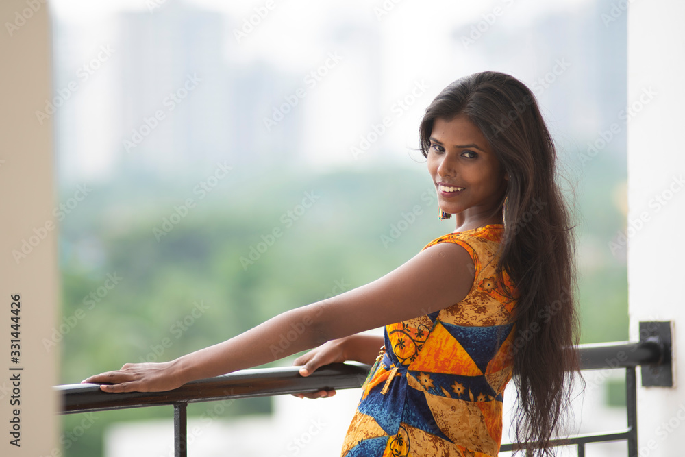 Φωτογραφία Fashion portrait of an young and beautiful brunette dark skinned Bengali woman in vibrant indo-western dress standing on a balcony in green urban background